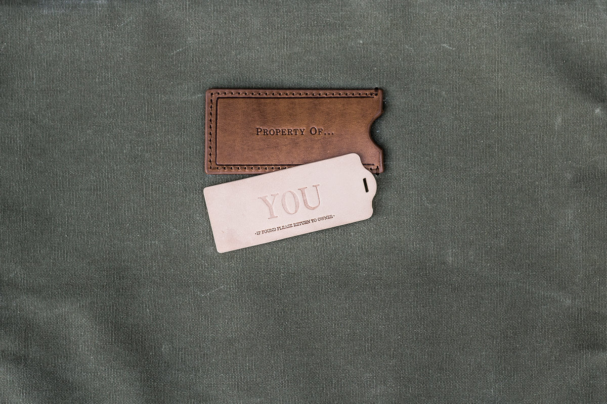 A brown leather Property Of... label on green fabric, next to a leather ID tag embossed with the word YOU
